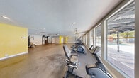 Located at 4870 Nellis Oasis Ln in Las Vegas, Nevada, this fitness center offers a generously sized, single-floor exercise room designed to accommodate a well-rounded workout experience. The open floor plan provides ample space for movement and equipment placement, with a durable concrete-style flooring surface throughout that is both practical and easy to maintain.

The facility is outfitted with a varied selection of cardio equipment, including treadmills, elliptical machines, and stationary bikes, offering users multiple options for aerobic training. A dedicated free weight area features a multi-tier dumbbell rack stocked with a full range of weights, while an additional strength and functional training station includes a cable machine, adjustable bench, and a multi-purpose rack with suspension training accessories.

Natural light fills the main exercise area through a bank of large windows that overlook an outdoor pool and covered patio area, creating a bright and motivating atmosphere. Recessed lighting and ceiling-mounted fan fixtures provide additional illumination and ventilation throughout the space. Safety features are present, including an AED unit, security camera, and clearly marked emergency exits. A thermostat and HVAC vents ensure climate control, an important comfort factor in the Las Vegas desert environment. The overall layout is functional, accessible, and well-suited for a community or shared fitness facility.