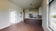 This single-floor unit showcases a clean, contemporary design with a cohesive neutral palette and high-quality finishes throughout. The open-concept kitchen is a standout feature, equipped with stainless steel appliances including a refrigerator, electric range, and over-the-range microwave. Crisp white upper cabinetry contrasts elegantly with dark wood-toned lower cabinets, complemented by light quartz countertops, a gray tile backsplash, and a matte black sink faucet. Pendant lighting and a linear under-cabinet light fixture add a modern touch to the space. Wood-look plank flooring flows seamlessly through the main living and kitchen areas, lending warmth and visual continuity. The entryway is practical and welcoming, featuring a built-in floating shelf and a keypad-enabled front door. The bedroom offers plush carpeting underfoot and a large window dressed with horizontal blinds, allowing generous natural light and outdoor views of surrounding greenery. The bathroom is finished with a modern floating vanity, a square undermount sink, matte black fixtures, and a framed mirror. A dedicated laundry area with a stacked washer and dryer provides convenient in-unit functionality. Overall, the space presents a well-appointed, move-in-ready layout with a cohesive modern aesthetic.