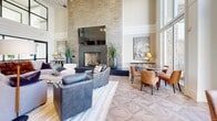 This elegantly designed single-floor communal space offers a seamless open-plan layout that integrates multiple distinct functional zones. The space is unified throughout by striking geometric parquet wood flooring, which adds warmth and visual interest underfoot.

The lounge area anchors one end of the space with a dramatic floor-to-ceiling stacked stone accent wall, serving as a backdrop for a large wall-mounted television and a sleek fireplace below. Comfortable seating arrangements, including plush sofas and accent chairs, create an inviting setting for relaxation and social gathering. Exposed wood beam detailing on the ceiling and an elegant chandelier contribute to the room's refined yet welcoming ambiance.

A long marble-topped kitchen island with leather bar stools runs through the heart of the space, illuminated by a row of industrial-style pendant lights. The adjacent full kitchen features white shaker cabinetry, dark stone countertops, a bold chevron-patterned tile backsplash, stainless steel appliances, and a built-in wine refrigerator — making it well-equipped for entertaining.

At the opposite end, floor-to-ceiling windows flood the space with abundant natural light and offer open views of the surrounding outdoor courtyard and community grounds. A casual dining area with round tables and leather chairs is positioned to take full advantage of these expansive views, creating a bright and airy atmosphere throughout.