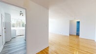 Located at 6001 Ch de la Côte-Saint-Luc in the borough of Hampstead, Québec, this well-maintained single-floor unit offers a thoughtfully laid-out living space comprising a living room, one bedroom, and a kitchen. Polished hardwood flooring runs throughout the living and sleeping areas, lending warmth and a clean, classic aesthetic to the interior. The living room is bright and open, featuring white walls, a front entry door, and a radiator heating unit along the wall, creating a simple and functional space. The bedroom is generously proportioned, offering ample room for furnishings, and includes a built-in closet with double doors. A gold multi-bulb ceiling fixture adds a subtle decorative touch. The kitchen is finished in an all-white palette, with white cabinetry, countertops, and full-size appliances including a refrigerator and electric stove. A window above the sink allows natural light to fill the space and provides a view of the surrounding greenery. The overall ambiance of the unit is clean, bright, and move-in ready, with a neutral color scheme that offers a versatile backdrop for personalization.