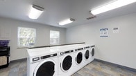 Located at 2414 Timbertrail Dr S in Columbus, Ohio, this single-floor community laundry facility is designed with functionality and convenience in mind. The room is outfitted with a row of commercial-grade Maytag front-loading washers and dryers, providing ample capacity to serve multiple users efficiently. A continuous countertop runs along the top of the machines, offering a practical surface for sorting and folding laundry. The space is finished with neutral gray walls and a durable tile floor, contributing to a clean and easy-to-maintain environment. Bright fluorescent ceiling fixtures ensure thorough, even illumination throughout the room, making it comfortable to use at any hour. Natural light also filters in through multiple windows fitted with blinds, adding to the welcoming atmosphere. Ventilation is addressed through ceiling vents, supporting a fresh and comfortable interior. Instructional signage is posted on the wall, providing users with clear guidance on proper machine use. Overall, the facility presents a tidy, well-organized space that prioritizes practicality and ease of use for its residents.