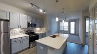 This single-story, one-bedroom, one-bathroom residence offers a thoughtfully designed open-concept layout that maximizes its compact footprint. The kitchen serves as the centerpiece of the home, showcasing gray shaker-style cabinetry paired with crisp white quartz countertops and a matching large kitchen island with an undermount sink and contemporary faucet. A stylish mosaic tile backsplash adds visual texture behind the stainless steel appliances, which include a range, microwave, and additional built-in units. Track lighting overhead ensures the kitchen is well-lit and functional.

Pendant lights with an industrial-chic aesthetic hang above the island, bridging the kitchen seamlessly into the living area beyond. The living space benefits from natural light streaming through large windows and a glass-paneled door that leads to an outdoor balcony, enhancing the sense of openness. A ceiling fan in the living area adds both comfort and style. Wood-look flooring flows throughout, contributing to a warm, cohesive aesthetic. The overall ambiance is modern and clean, with neutral tones creating a versatile, move-in-ready environment.