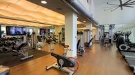 This fitness center offers a spacious, single-floor layout designed to accommodate a wide variety of workout routines. The space is thoughtfully organized, with cardio equipment — including LifeCycle stationary bikes and treadmills — positioned alongside a selection of strength training machines from Life Fitness and similar brands. The open floor plan allows for comfortable movement between stations without feeling crowded.

Architecturally, the space features a distinctive sculpted ceiling with wave-like panels in one section, adding a modern aesthetic contrast to the exposed industrial-style ductwork visible in another area. Structural columns are integrated into the layout, subtly dividing the space into functional zones while maintaining an overall open feel. The warm hardwood flooring runs throughout, lending a clean, polished look to the environment.

Large mirrors are strategically placed to enhance the sense of space and allow users to monitor their form during workouts. Recessed lighting provides consistent, even illumination throughout. The overall ambiance is modern, clean, and professional, creating an inviting environment well-suited for focused exercise.