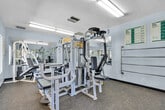 Located at 1401 E Santo Antonio Dr in Colton, California, this dedicated fitness center offers a well-equipped exercise space on a single floor. The room is designed to maximize functionality, housing multiple cable and strength training machines, including a multi-station cable system, a lat pulldown unit, and various benches suited for a range of resistance exercises. Durable speckled rubber flooring throughout provides stability and easy maintenance underfoot. Large wall-mounted mirrors line two sides of the room, creating a sense of expanded space and allowing users to monitor their form during workouts. Fluorescent overhead lighting ensures the room is consistently well-lit. Windows along one wall invite natural light and offer a direct view of an outdoor pool area, adding a refreshing visual element to the workout environment. Informational workout and exercise guide charts are posted on the white walls, offering convenient reference material for users. A horizontal towel or stretching bar is also mounted to the wall, adding a practical touch. The space is accessed through a single door and is efficiently arranged to accommodate multiple users simultaneously despite its compact footprint.