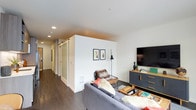 This single-floor residence offers a thoughtfully designed living space with a contemporary aesthetic. The living area serves as the central hub, featuring rich dark hardwood flooring that runs throughout, creating a warm contrast against the crisp white walls and smooth white ceiling with recessed lighting. A wall-mounted flat-screen television is positioned above a stylish charcoal and wood media console, while a cozy seating arrangement anchors the space. A statement floor lamp and curated wall art add personality to the room.

A defining architectural feature is the frosted glass barn door, which slides along a mounted track to provide privacy while maintaining an open, airy feel. This detail adds both functional and visual interest to the layout. Beyond the living area, a hallway extends toward additional rooms, hinting at a well-organized floor plan. The frosted glass panels allow natural light to filter through, contributing to the overall brightness of the space.

The interior design balances modern minimalism with warm textures through layered soft furnishings and natural materials, resulting in a space that feels both polished and livable.