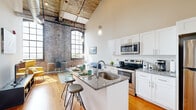 This loft-style unit masterfully combines industrial character with contemporary design. The open-plan kitchen and living area is the architectural centerpiece, boasting dramatically high ceilings with exposed original timber beams and expansive arched, grid-pane windows that flood the space with natural light. Original exposed brick walls add raw, historic texture throughout, contrasting beautifully with the clean, modern finishes.

The kitchen is well-appointed with white shaker cabinetry, granite countertops, a subway tile backsplash, stainless steel appliances, and a peninsula island with built-in sink and bar seating — ideal for both cooking and casual dining. Warm hardwood floors run throughout the main living areas, enhancing the inviting ambiance.

The living area benefits from the same soaring ceilings and striking brick wall backdrop, with generous natural light streaming through large industrial-style windows offering open views.

The bedroom is a quieter retreat, featuring carpet flooring, tall ceilings, a sliding-door closet, and an exposed brick accent wall with its own large window. The bathroom offers a tiled tub-shower combo with marble-style tile surround, granite vanity, and white cabinetry. An in-unit laundry completes this efficiently designed space.