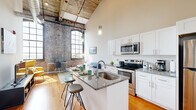 This loft-style unit masterfully combines industrial character with contemporary design. The open-plan kitchen and living area is the architectural centerpiece, boasting dramatically high ceilings with exposed original timber beams and expansive arched, grid-pane windows that flood the space with natural light. Original exposed brick walls add raw, historic texture throughout, contrasting beautifully with the clean, modern finishes.

The kitchen is well-appointed with white shaker cabinetry, granite countertops, a subway tile backsplash, stainless steel appliances, and a peninsula island with built-in sink and bar seating — ideal for both cooking and casual dining. Warm hardwood floors run throughout the main living areas, enhancing the inviting ambiance.

The living area benefits from the same soaring ceilings and striking brick wall backdrop, with generous natural light streaming through large industrial-style windows offering open views.

The bedroom is a quieter retreat, featuring carpet flooring, tall ceilings, a sliding-door closet, and an exposed brick accent wall with its own large window. The bathroom offers a tiled tub-shower combo with marble-style tile surround, granite vanity, and white cabinetry. An in-unit laundry completes this efficiently designed space.