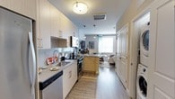 This studio offers a thoughtfully designed open-concept layout that seamlessly integrates the kitchen, dining, and living areas into one cohesive space. The modern kitchen is a standout feature, showcasing crisp white cabinetry with gold-toned hardware, a subway tile backsplash, dark countertops, and stainless steel appliances including a dishwasher and range. A peninsula island with pendant lighting serves as a natural dividing point between the kitchen and living area, providing additional prep space and a casual dining option. Light wood-tone flooring runs throughout, adding warmth and visual continuity. The living area is bathed in natural light from a large window dressed with teal curtains, creating a bright and welcoming ambiance. A flush-mount ceiling fixture illuminates the kitchen corridor, while pendant lights above the island add a contemporary touch. A closet with double doors provides practical storage solutions. The bathroom completes this unit, offering a full private bath. The overall aesthetic is clean, modern, and functional, making this studio an efficient and stylish living space suited for everyday comfort.