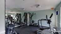 This fitness center is thoughtfully designed to accommodate a comprehensive workout experience. The space is finished with soft sage-green walls accented by white trim, creating a calm and focused atmosphere conducive to exercise. A large mirrored wall spans one side of the room, visually expanding the space and allowing users to monitor their form during workouts. The flooring consists of durable dark rubber matting, providing both safety and comfort underfoot. Overhead, flush-mount ceiling lights offer consistent, even illumination throughout the room, complemented by a ceiling-mounted ventilation unit that ensures proper airflow. The facility is outfitted with a multi-station cable and weight machine, offering a wide range of strength training exercises, alongside additional resistance and functional training equipment. Cardio offerings include a True-brand elliptical trainer, delivering a low-impact aerobic option for users. An instructional exercise chart is posted on the wall for easy reference. The overall layout is organized and efficient, making effective use of the available floor space while keeping pathways clear between machines. This single-floor fitness center is a well-maintained, fully functional amenity suited for a variety of fitness levels and training goals.