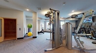 This fitness center is thoughtfully designed to accommodate a wide range of workout styles across several distinct zones. The cardio area features a row of treadmills, ellipticals, and stair climbers lined against a darkly textured accent wall, with wall-mounted televisions providing entertainment during workouts. The flooring transitions between patterned carpet and herringbone-style tile, adding visual variety throughout the space.

The strength training section is outfitted with professional-grade Hoist cable machines, a full dumbbell rack ranging across multiple weights, an adjustable workout bench, and an orange rubberized lifting platform. A large circular mirror reflects the equipment area, enhancing the sense of openness. A functional training zone includes stability balls in assorted sizes, medicine balls on a storage rack, suspension trainers, and a rowing machine, offering versatile options for conditioning and flexibility work.

Natural light enters through large windows offering outdoor views, contributing to an airy atmosphere. Recessed lighting throughout ensures consistent illumination. A standout feature is a cedar-paneled sauna visible in the overview, providing a dedicated recovery space. Artwork on the walls and clean, neutral tones complete the polished, modern ambiance of this comprehensive fitness facility.