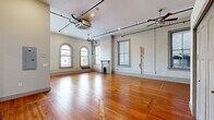 This single-floor unit masterfully combines historic architectural charm with contemporary upgrades. Rich, original hardwood floors run throughout the entire space, grounding each room with warmth and character. The expansive open living area commands attention with its soaring ceilings, exposed industrial-style conduit, ceiling fans, and a row of elegant arched windows that flood the room with natural light and street views. A decorative fireplace with a classic white mantle serves as a refined focal point within the living space, adding a timeless touch to the interior.

The kitchen has been thoughtfully renovated and features white shaker cabinetry, granite countertops, stainless steel appliances, and a generous island with a built-in sink — all illuminated by a statement pendant light and framed by the same graceful arched windows. The layout is both functional and visually striking.

The bedroom offers a calm, well-lit retreat with a large closet, ceiling fan, and a window with natural light. The bathroom is clean and modern, appointed with a full tub and shower combination and a dark-stained vanity with a white countertop, all tied together by the consistent warm hardwood flooring. This space is a compelling blend of historic fabric and updated living.
