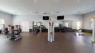 This fitness center is a spacious, single-floor facility designed to accommodate a comprehensive range of workout routines. The interior is finished with light gray walls accented by a dark wainscoting band, recessed lighting throughout, and durable speckled rubber flooring that provides both safety and resilience underfoot.

The cardio zone features a row of True elliptical trainers and treadmills positioned along one wall, allowing users to exercise while benefiting from natural light streaming through multiple windows fitted with blinds. Opposite this area, a dedicated strength-training section houses a multi-functional cable and resistance machine, along with additional selectorized equipment suited for targeted muscle training.

A free weights area includes an adjustable workout bench, dumbbells organized on a rack, and a large wall-mounted mirror that enhances the sense of space and supports proper form during exercise. Mounted flat-screen televisions are positioned for visibility from multiple workout stations throughout the room.

A distinct functional training zone, finished with a yellow-green floor accent, features TRX suspension trainers mounted to the wall alongside open floor space for dynamic movements. The overall layout is thoughtfully organized to separate cardio, strength, and functional training areas, creating a practical and efficient environment for users of varying fitness levels.