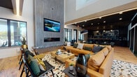 This thoughtfully designed single-floor communal lounge space offers a sophisticated blend of comfort, style, and functionality. The centerpiece of the main lounge area is a striking floor-to-ceiling stone-clad fireplace wall, fitted with a wide-format flat-screen television and a sleek linear fireplace below — together creating a dramatic focal point that draws the eye upward toward the soaring double-height ceiling. Generous amber leather sectional sofas, patterned cowhide rugs, and rustic wood coffee tables furnish the lounge, lending a warm yet contemporary character. Natural light filters through large black-framed windows, offering views to an outdoor terrace area. Adjacent to the main lounge, a dedicated café-style bar and kitchen zone features dark cabinetry, a richly veined marble backsplash, pendant lighting, a coffee station, and a large communal high-top table beneath a distinctive slatted wood ceiling — ideal for casual gatherings or informal meetings. A separate co-working nook provides round dining-height tables and workstation seating, complemented by curated artwork and accent lighting. Throughout, warm hardwood flooring, linear LED cove lighting, and bold dark accent walls tie the spaces together into a cohesive, upscale aesthetic.