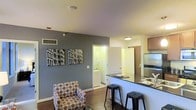 This single-floor unit is designed with an open-concept layout that seamlessly connects the living area and kitchen. The living space features rich hardwood-style flooring and a neutral gray accent wall, adding depth and a modern aesthetic to the room. Decorative wall art depicting urban architecture enhances the contemporary feel of the space.

The kitchen is positioned along one side of the open floor plan, defined by a dark-topped breakfast bar with bar stool seating — an efficient use of space that also serves as a casual dining area. The kitchen is equipped with stainless steel appliances, including a refrigerator, and features warm wood-toned cabinetry overhead. A sink is visible along the counter, contributing to a fully functional cooking space.

A hallway visible from the main living area leads to a dedicated in-unit laundry area, with a washer and dryer clearly visible, offering added convenience. The unit benefits from flush-mount ceiling lighting that provides even illumination throughout. The overall ambiance is modern and functional, with a clean, well-organized layout suited for everyday living.