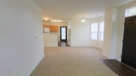 Located at 100 Reserve Ct in Glenville, NY, this single-floor residence offers a well-designed, open-concept layout that promotes a sense of spaciousness and easy flow between living and dining areas. The living room features neutral-toned carpeting and clean white walls, creating a bright and welcoming atmosphere. A pendant light fixture adds a decorative touch overhead, while a large window allows natural light to fill the space.

The kitchen is positioned adjacent to the living area and is outfitted with warm wood-toned cabinetry, a white microwave, a full-size refrigerator, and a light-colored countertop that extends to form a convenient breakfast bar or pass-through. A contrasting dark-framed glass panel door provides both a design accent and direct access to the exterior, adding functionality alongside visual interest.

The overall color palette throughout the space is kept light and neutral, with white walls and ceilings that enhance brightness and complement the natural materials used in the cabinetry. The open arrangement between the kitchen and living area makes the space feel cohesive and practical, well-suited for everyday living and entertaining alike.