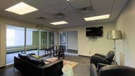 This single-floor commercial space is designed as a functional and welcoming lounge or common area, offering a well-organized layout suited for relaxation, informal gatherings, or casual meetings. The room features a drop ceiling with recessed fluorescent lighting panels that provide consistent, even illumination throughout the space.

Large glass partition walls and glass-paned doors form a defining architectural element, separating interior zones while maintaining an open, connected feel and allowing natural light to flow freely between areas. The walls are finished in a neutral cream tone, contributing to a clean and professional atmosphere.

Seating is arranged thoughtfully, with black leather sofas and chairs positioned around a patterned area rug, creating a comfortable lounging zone. A high-top table with accompanying bar stools offers an alternative seating option for those preferring an upright setting. A wall-mounted flat-screen television serves as a focal point on one wall, adding a media-friendly element to the space.

The flooring consists of commercial-grade carpet tiles, practical and durable for high-traffic use. Visible HVAC vents in the ceiling ensure proper climate control throughout. Overall, the space strikes a balance between comfort and functionality, making it well-suited for a community room, waiting area, or collaborative lounge environment.