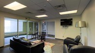 This single-floor commercial space is designed as a functional and welcoming lounge or common area, offering a well-organized layout suited for relaxation, informal gatherings, or casual meetings. The room features a drop ceiling with recessed fluorescent lighting panels that provide consistent, even illumination throughout the space.

Large glass partition walls and glass-paned doors form a defining architectural element, separating interior zones while maintaining an open, connected feel and allowing natural light to flow freely between areas. The walls are finished in a neutral cream tone, contributing to a clean and professional atmosphere.

Seating is arranged thoughtfully, with black leather sofas and chairs positioned around a patterned area rug, creating a comfortable lounging zone. A high-top table with accompanying bar stools offers an alternative seating option for those preferring an upright setting. A wall-mounted flat-screen television serves as a focal point on one wall, adding a media-friendly element to the space.

The flooring consists of commercial-grade carpet tiles, practical and durable for high-traffic use. Visible HVAC vents in the ceiling ensure proper climate control throughout. Overall, the space strikes a balance between comfort and functionality, making it well-suited for a community room, waiting area, or collaborative lounge environment.