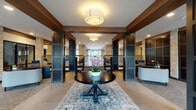 This thoughtfully designed single-floor communal space combines contemporary and transitional architectural elements to create an inviting, upscale environment. The grand entry foyer sets the tone immediately, anchored by a dark-stained round pedestal table topped with fresh florals, positioned atop a layered area rug over large-format tile flooring. Dramatic dark-paneled columns contrast beautifully with stacked natural stone pillars throughout, while warm wood beam accents frame the ceiling and define distinct zones within the open layout.

Drum pendant fixtures provide ambient illumination across the entry and adjoining corridors, where carpeted hallways lead to additional rooms flanked by stone-clad columns. A spacious lounge area features light hardwood flooring, a teal-accented fireplace wall with built-in shelving, and eclectic furnishings including vibrant orange accent chairs, a plush sectional, and bold patterned rugs. A decorative globe chandelier serves as a focal point overhead.

An upper-level overlook with sleek metal railings is visible from the main floor, emphasizing the volume and vertical scale of the space. Floor-to-ceiling sheer-draped windows flood the lounge with natural light, while stacked stone walls add warmth and texture throughout. The overall ambiance is polished, welcoming, and carefully curated.
