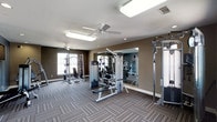 This fitness center offers a dedicated space for strength training and exercise, thoughtfully designed to accommodate a variety of workout routines. The room features neutral, earth-toned walls that create a focused and energizing atmosphere, complemented by patterned, low-pile carpet flooring that provides both comfort and durability underfoot. Ceiling fans are installed overhead to maintain airflow throughout the space, contributing to a comfortable workout environment alongside recessed and panel lighting that ensures the area is well-lit at all times. A large mirrored wall along one side of the room expands the sense of space while allowing users to monitor their form during exercise. The fitness center is outfitted with a selection of strength training machines, including cable and pulley systems, multi-function weight stations, and adjustable benches, offering options for both upper and lower body workouts. Natural light filters in through windows along the perimeter wall, adding a bright and open feel to the interior. The overall layout is practical and spacious, providing ample room to move between equipment, making this fitness center a functional and well-appointed amenity.