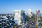 Located along the iconic Ocean Boulevard in Long Beach, California, this modern 2-bedroom, 2-bathroom residence offers a thoughtfully designed open-concept layout with clean contemporary finishes throughout. Light hardwood flooring flows seamlessly across the living spaces, complementing the bright, white walls and creating a cohesive, airy ambiance.

The open kitchen and living area is a standout feature, anchored by a sleek, well-appointed kitchen with two-tone cabinetry — white upper cabinets paired with warm natural wood lower cabinets — set against white quartz countertops. A full suite of stainless steel appliances includes a gas range, built-in microwave, dishwasher, and French door refrigerator. The living area opens to expansive floor-to-ceiling windows and sliding glass doors that frame striking panoramic views of the Long Beach skyline and surrounding cityscape, flooding the space with natural light.

Both bedrooms are well-proportioned and bright, each benefiting from large windows with urban views. The primary bedroom includes access to a dedicated closet. The two bathrooms feature light wood vanity cabinetry, white countertops, large-format tile flooring, and glass-enclosed walk-in showers, maintaining the clean, modern aesthetic found throughout the residence.