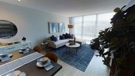 This single-floor space offers a thoughtfully designed open-concept layout that seamlessly connects the dining and living areas, creating a cohesive and functional environment. The living room is anchored by a large sectional sofa dressed with navy and neutral-toned accent pillows, complemented by a round marble-top coffee table and a bold navy geometric area rug that adds depth and character to the space. A striking floor lamp provides warm ambient lighting, while a pair of coordinating blue abstract artworks adorns the walls, reinforcing the space's refined color palette. The dining area features a dark-toned table paired with leather accent chairs, offering an elegant setting for everyday meals or entertaining. Floor-to-ceiling windows with vertical blinds flood the interior with abundant natural light and create an airy, expansive atmosphere. Light hardwood-style flooring runs throughout, enhancing the bright and contemporary feel of the space. Lush indoor greenery adds a touch of warmth and life to the overall aesthetic. The interior design balances modern minimalism with inviting comfort, making it both stylish and livable.