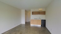 This single-floor unit presents a clean, minimalist design characterized by polished concrete floors and smooth, neutral-toned walls that create a bright and functional atmosphere. The open-plan layout integrates the living area and kitchenette into one cohesive space, maximizing usability.

The kitchenette is positioned along one wall and features light maple wood cabinetry with a compact countertop, a built-in sink, and an electric cooktop. Open wooden shelving mounted above the cabinetry provides additional storage and display space. A compact black mini-refrigerator sits alongside the lower cabinets, completing the kitchen setup efficiently within a small footprint.

The main living area is open and flexible, with polished concrete flooring throughout that adds an industrial character to the otherwise simple interior. A partial wall division near one corner creates a semi-private alcove housing a closet with hanging space, suggesting a sleeping or personal area separate from the main room.

The textured white ceiling and neutral wall tones keep the space feeling light and airy. The layout is straightforward and well-suited for a variety of uses, offering a functional, no-frills environment.