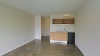 This single-floor unit presents a clean, minimalist design characterized by polished concrete floors and smooth, neutral-toned walls that create a bright and functional atmosphere. The open-plan layout integrates the living area and kitchenette into one cohesive space, maximizing usability.

The kitchenette is positioned along one wall and features light maple wood cabinetry with a compact countertop, a built-in sink, and an electric cooktop. Open wooden shelving mounted above the cabinetry provides additional storage and display space. A compact black mini-refrigerator sits alongside the lower cabinets, completing the kitchen setup efficiently within a small footprint.

The main living area is open and flexible, with polished concrete flooring throughout that adds an industrial character to the otherwise simple interior. A partial wall division near one corner creates a semi-private alcove housing a closet with hanging space, suggesting a sleeping or personal area separate from the main room.

The textured white ceiling and neutral wall tones keep the space feeling light and airy. The layout is straightforward and well-suited for a variety of uses, offering a functional, no-frills environment.