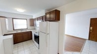 This single-floor space offers a practical and well-organized layout designed for comfortable everyday living. The kitchen is a standout feature, showcasing rich dark wood cabinetry that lines the walls from countertop to ceiling, providing generous storage throughout. The kitchen is equipped with a white refrigerator, an electric range with oven, and a built-in sink beneath a window that allows natural light to brighten the workspace. Light-toned tile flooring runs throughout the kitchen, creating a clean and easy-to-maintain surface. A transitional opening connects the kitchen directly to an adjoining room, which features warm hardwood flooring that contrasts with the tile and adds a sense of warmth to the overall space. The open sightline between the two areas enhances the flow and makes the layout feel cohesive and connected. The neutral wall tones throughout contribute to a bright and airy atmosphere, offering a versatile backdrop suitable for a variety of interior styles. Overall, the space presents a straightforward, functional design that balances utility with a welcoming ambiance.
