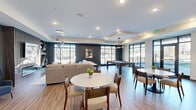 This contemporary amenity space spans a generous single floor, thoughtfully designed to offer a variety of recreational and social experiences. The entrance features floor-to-ceiling glass panels and black-framed doors that flood the foyer with natural light and offer views of the surrounding landscape.

The main clubroom is an open, inviting area with warm wood-look flooring, tray ceilings, and large windows that frame outdoor views. A statement fireplace clad in marble-style tile anchors the lounge seating area, complemented by a slatted wood accent wall and pendant globe lighting. Comfortable sofas and accent chairs create an intimate gathering atmosphere.

Adjacent to the lounge, a full bar kitchen impresses with a long quartz countertop island, patterned tile bar seating, backlit open shelving lined with glassware and greenery, a sleek tile backsplash, and a built-in media screen — ideal for entertaining.

A billiards area with a full-size pool table sits beneath modern globe pendant lights, surrounded by floor-to-ceiling windows that invite the outdoors in. A separate golf simulator room offers a cozy lounge sofa, framed golf artwork, a large display screen, and a dedicated club rack — a standout amenity for enthusiasts. The space as a whole delivers a polished, upscale ambiance suited for both casual relaxation and lively social gatherings.
