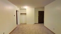 This single-floor space presents a straightforward and functional layout with clean, neutral-toned walls throughout. The main room is finished with wall-to-wall carpeting in a light beige tone, providing a soft underfoot feel and contributing to the overall warmth of the interior. Ceiling-mounted lighting illuminates the space adequately, keeping the atmosphere bright and even.

A prominent open closet is built into the wall, featuring a single hanging rod and a shelf above, offering practical storage without the need for additional furniture. The closet opening is framed cleanly into the wall, maintaining a tidy appearance. Adjacent to the closet, a solid dark wood entry door serves as the main point of access, fitted with a standard lock mechanism. The door's darker tone provides a visual contrast against the light-colored walls.

A secondary doorway is visible, suggesting additional rooms or utility spaces beyond the main area. Baseboards run along the perimeter of the room, adding a modest finishing detail to the overall design. The space is compact yet organized, with a simple, no-frills aesthetic that prioritizes utility and ease of use.