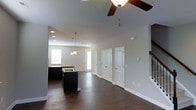 This two-story space showcases a well-designed open-concept main floor that seamlessly connects the kitchen and living areas. The kitchen features dark cabinetry paired with a contrasting light countertop, an undermount sink, and a central island that provides additional workspace and storage. Pendant lighting hangs above, complementing the recessed lighting throughout the ceiling. A ceiling fan with a warm-toned light fixture anchors the living area, contributing to both comfort and style.

Rich, dark hardwood-style flooring runs throughout the main level, providing a striking contrast against the neutral gray-toned walls and crisp white trim and door casings. Multiple interior doors with classic panel detailing line the far wall, leading to additional rooms or utility spaces. Natural light filters in through windows on either side of the space, brightening the open layout.

A staircase is visible toward the rear, indicating access to the second floor. The clean lines, neutral palette, and thoughtful lighting choices throughout the main floor create a cohesive, modern aesthetic that balances functionality with visual appeal.
