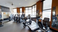 This fitness center is a thoughtfully designed workout facility that combines functionality with an inspiring atmosphere. The space is dominated by a generous row of premium cardio equipment, including elliptical trainers, stationary bikes, and treadmills, all outfitted with individual digital display screens for an engaging exercise experience. Warm-toned accent walls in rich burnt orange complement the neutral flooring, creating an energetic yet focused environment. The most striking architectural feature is the expansive floor-to-ceiling window system that wraps along the perimeter of the space, flooding the interior with abundant natural light and offering sweeping panoramic views of the surrounding cityscape and beyond. Recessed ceiling lighting supplements the natural light, ensuring the space is well-lit at any hour. The open layout allows for comfortable movement between machines, and the overall design strikes a balance between a professional gym setting and an elevated, amenity-style experience. The combination of high-end equipment, thoughtful design details, and dramatic exterior views makes this fitness center a standout facility suited for a rigorous and motivating daily workout routine.