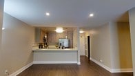 This single-floor space offers a thoughtfully designed open-concept layout that seamlessly connects the kitchen and living areas, creating a spacious and airy feel. The kitchen is a standout feature, showcasing contemporary light wood cabinetry, a clean white countertop peninsula that doubles as a breakfast bar, and stainless steel appliances including a full-size refrigerator and microwave. Pendant lighting with warm-toned bulbs hangs above the peninsula, complementing the recessed ceiling lights that illuminate the space evenly throughout. The neutral beige walls and crisp white trim provide a clean, versatile backdrop that suits a variety of décor styles. Rich, dark hardwood-style flooring runs continuously through the open area, grounding the space with warmth and visual cohesion. A hallway visible from the main living area suggests additional rooms beyond, hinting at a well-organized floor plan. The overall ambiance is modern, clean, and inviting — ideal for everyday living and casual entertaining alike.