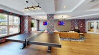 Located at 8 Norden Place in Norwalk, CT, this single-floor amenity space is a lively and thoughtfully designed recreational lounge that seamlessly blends entertainment and social gathering. The room is defined by its bold, large-scale botanical wallcoverings in rich coral and teal tones, which are carried across accent walls and structural columns, lending the space a cohesive and energetic personality.

The game area is generously sized to accommodate multiple pool tables and a ping-pong table, providing ample room for recreational play. Warm hardwood-style flooring flows throughout, grounding the vibrant decor with a natural, inviting feel. Coffered ceilings with recessed lighting add architectural interest and height, while a series of modern pendant and sputnik-style chandeliers provide layered, stylish illumination.

A sleek bar counter finished in light stone tops a slatted base panel, lined with a row of navy velvet barstools — ideal for socializing or casual dining. Mounted flat-screen TVs are positioned throughout the room for entertainment viewing. The lounge seating zone features plush caramel leather sofas atop a graphic blue area rug, arranged around low coffee tables for a relaxed atmosphere.

Large glass doors and wide windows flood the space with natural light and offer direct access to an outdoor patio area, seamlessly connecting interior comfort with the outdoors.