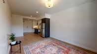 This well-designed single-floor unit offers a clean, contemporary layout with light hardwood flooring running throughout every room, creating a cohesive and warm aesthetic. The open-concept living and kitchen area is efficiently arranged, with the kitchen featuring dark wood cabinetry, stainless steel appliances including a full-size refrigerator, a single-basin sink, open shelving for storage, and a dishwasher. Recessed lighting and a flush-mount ceiling fixture keep the space bright and functional. The entry area is well-defined, with a solid front door and a clear sightline into the living space.

The bedroom is a standout area, benefiting from a large window that frames a picturesque view of surrounding greenery and neighboring architecture, filling the room with natural light. A wall-mounted HVAC unit provides climate control, and a sliding-door closet offers practical storage. An additional room provides further flexible living or storage space.

The bathroom is neatly appointed with a dark wood vanity, a single sink with a light countertop, a large mirror with overhead lighting, and hardwood-style flooring that ties into the rest of the unit. In-unit laundry adds everyday convenience. Overall, the unit presents a modern, functional, and well-lit living environment.