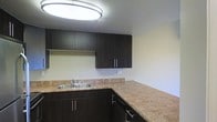 This single-floor space showcases a kitchen designed with a clean, contemporary aesthetic. The kitchen is appointed with dark espresso-toned cabinetry, featuring sleek bar-pull hardware that adds a modern touch. The upper and lower cabinets provide ample storage, complementing the generous counter space finished in warm granite-style laminate countertops that extend in an L-shaped configuration, offering excellent workspace for meal preparation. A double-basin stainless steel sink is integrated into the countertop, paired with a standard faucet fixture. The kitchen is well-lit by a large circular flush-mount ceiling light that provides bright, even illumination throughout the space. The light-colored walls contrast effectively with the dark cabinetry, creating a balanced visual dynamic. A dishwasher is discreetly integrated beneath the countertop, adding functionality. The overall design of the kitchen emphasizes practicality and modern style, with a neutral color palette that offers versatility for a range of interior design preferences.