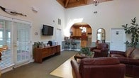 This single-floor space makes a strong impression with its dramatically vaulted ceilings accented by exposed wood beam detailing, creating an open and airy atmosphere throughout. The main lounge area is furnished with rich, dark leather seating arranged around a wooden coffee table, offering a welcoming and comfortable gathering space. A wall-mounted flat-screen television is positioned above a wooden media console, which serves as a functional focal point for the room. Natural light filters in from above, complementing the warm tones of the wood trim and neutral wall colors. An elegant arched passageway connects the lounge to a well-appointed kitchen area, which features wooden cabinetry, modern appliances including a refrigerator and microwave, and bar-height seating with decorative wrought iron stools. A chandelier overhead adds a refined touch to the kitchen space. Decorative greenery and thoughtful accent pieces are placed throughout, lending a homey and lived-in warmth to the environment. A mirror near the entry reflects additional light and adds visual depth. The overall ambiance blends comfort with tasteful design, making this a welcoming and functional space suited for everyday use and shared living.