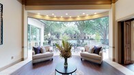 This single-story lounge area is defined by its striking curved wall of floor-to-ceiling glass windows, which serve as the architectural centerpiece of the space. The panoramic glazing floods the interior with abundant natural light and provides unobstructed views of a landscaped outdoor area featuring a pool, patio seating, and mature trees that create a lush, green backdrop.

The interior is finished with rich hardwood-style flooring that adds warmth and continuity throughout the space. A curved tray ceiling with recessed lighting follows the arc of the window wall, enhancing the room's architectural character and providing soft, even illumination that complements the natural light.

The seating arrangement consists of two neutral-toned linen armchairs accented with navy and patterned throw pillows, positioned symmetrically around a small, round dark-topped coffee table. A large floral arrangement serves as a focal centerpiece, adding color and an organic element to the refined setting.

Overall, the space strikes a balance between elegance and openness, with its seamless indoor-outdoor connection and thoughtfully curated furnishings creating a welcoming and sophisticated atmosphere.