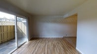 This single-floor space presents a clean, minimalist aesthetic centered around light-toned hardwood flooring that runs throughout the main living area. The flooring transitions to tile in an adjacent section, suggesting a distinct functional zone nearby. Neutral, light gray walls provide a versatile backdrop that complements the overall brightness of the room. A large sliding glass door along one wall serves as a prominent architectural feature, flooding the interior with natural light and offering a direct view to an outdoor area, likely a patio or balcony. The door's generous dimensions allow sunlight to cast dynamic patterns across the floor, enhancing the open, airy feel of the space. White baseboards provide a crisp contrast against both the walls and flooring, adding a polished finish to the room's clean lines. The ceiling is smooth and painted in a soft neutral tone, contributing to the room's cohesive, understated palette. The layout appears open and unobstructed, making the space adaptable for a variety of furniture arrangements. Overall, the space conveys a practical yet comfortable ambiance suited for everyday living.