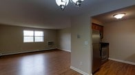 This single-floor space offers a straightforward and functional layout, connecting an open living area to a kitchen. The living area is finished with warm hardwood-style flooring that extends throughout, complemented by neutral beige walls and clean white baseboards that create a bright, understated aesthetic. A classic multi-arm chandelier serves as a central lighting fixture, adding a traditional touch to the space. A window along the exterior wall allows natural light to filter in, while a wall-mounted HVAC unit and baseboard heating provide year-round climate control. The kitchen is partially visible through an open passageway, where rich cherry-toned upper cabinetry contrasts with stainless steel appliances, including a full-size refrigerator and range. The transition between spaces is defined by a structural column, giving each area a sense of separation while maintaining an open feel. The overall ambiance is clean and practical, with a neutral palette that provides a versatile backdrop well-suited for a variety of interior styles.