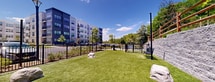 Located at 6 Becker Farm Rd in Roseland, NJ, this outdoor dog park is a well-designed amenity space serving the adjacent multi-story residential apartment community. The park features a generously sized, level ground surface covered in vibrant artificial turf, providing a clean and low-maintenance area for pets to roam freely. Scattered throughout the space are several natural boulder accents that add a rugged, organic aesthetic to the otherwise manicured grounds. The park is fully enclosed by sturdy black metal fencing, ensuring the safety and containment of pets at all times. A dedicated entry gate provides controlled access, and pet waste stations are conveniently positioned throughout the area to promote cleanliness and responsible pet ownership. Along one perimeter, a textured granite block retaining wall is topped by a warm-toned wooden rail fence, creating a defined boundary that also serves as an attractive landscape feature. Mature trees line the backdrop, offering natural shade and a sense of seclusion. The surrounding modern apartment buildings, characterized by bold navy and light gray exteriors with large windows, are clearly visible, reinforcing this space's role as a premium community amenity. Ample open sky above ensures plenty of natural light throughout the day.