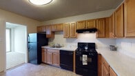 This single-floor residence offers a practical and comfortable layout across several well-defined spaces. The kitchen is equipped with warm honey-toned wood cabinetry providing generous upper and lower storage, light speckled countertops, and a full suite of black appliances including a gas range, dishwasher, and refrigerator. A ventilation hood is mounted above the stove, and tile-style flooring grounds the workspace.

The living area is expansive and filled with natural light from a wide bank of floor-to-ceiling windows dressed with vertical blinds, offering views of the surrounding urban skyline. A baseboard heating unit runs along the window wall, and neutral carpet extends throughout. Textured ceilings are consistent across the space.

The bedroom is generously sized with soft carpeting, access to a walk-in closet fitted with a hanging rod, and a connecting hallway leading to the bathroom. The bathroom features a wide vanity with a large mirror, ample under-sink storage with white cabinetry, light countertops, and tile flooring. The overall palette throughout is neutral and understated, lending a clean, move-in-ready ambiance.