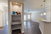 This single-story residence offers a well-organized layout with a cohesive neutral color palette throughout. Dark hardwood-style flooring spans the main living areas, complemented by crisp white trim, crown molding, and ceiling fans. The living area is spacious and open, receiving natural light through large windows, and connects seamlessly to the dining and kitchen spaces.

The kitchen is well-appointed with white cabinetry, granite countertops, a dark subway tile backsplash, stainless steel appliances including a refrigerator, range, and dishwasher, and pendant lighting above a breakfast bar peninsula. The open-concept design makes it ideal for both cooking and entertaining.

A distinctive built-in nook near the entry features a granite-topped desk surface and an open display shelf above, providing a practical workspace or organizational area. A louvered door nearby leads to a utility or storage closet.

The bedrooms are carpeted for comfort, each featuring ceiling fans and windows that allow natural light. At least one bedroom includes an accessible walk-in closet. The bathroom includes a granite-topped vanity with an undermount sink and ample counter space. Overall, the space presents a clean, functional, and modern interior.