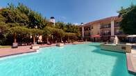 This amenity space combines a stunning outdoor pool area with a well-appointed fitness center, delivering a full-featured recreational experience. The expansive pool features crystal-clear turquoise water and is framed by a generous pool deck lined with lounge chairs, ideal for relaxing under open skies. Mature trees and lush greenery surround the area, providing natural shade and a serene atmosphere, while a pergola-style structure adds architectural charm to the outdoor setting.

The fitness center is thoughtfully organized across distinct workout zones, all unified by stylish herringbone-patterned carpet flooring. The main strength area is equipped with a multi-functional cable and power rack system, a full dumbbell rack, medicine balls, battle ropes, and stability balls, catering to a wide range of training styles. A separate cardio zone features treadmills positioned beneath a wall-mounted TV, bathed in natural light from large windows overlooking the surrounding grounds. Additional strength equipment includes a rowing machine, cable machine, squat rack, and bench press station. The fitness center benefits from high ceilings, recessed lighting, a ceiling fan, and large windows throughout, creating a bright and airy environment. A refreshment and towel station adds a convenient finishing touch.