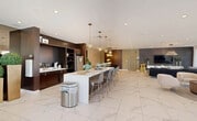 Located in Princeton, FL, this single-story community clubhouse offers a generously sized, open-plan gathering space designed with a contemporary aesthetic throughout. The interiors are unified by large-format marble-look porcelain tile flooring that reflects an abundance of natural light, creating a bright and welcoming atmosphere.

The kitchen area serves as a central hub, anchored by a long white waterfall island with seating for multiple guests. Dark-toned cabinetry with open shelving lines the walls, complemented by stainless steel appliances and globe pendant lighting in warm brass tones. Adjacent to the kitchen, a formal dining area features an elegant wood-grain conference-style table surrounded by plush navy velvet chairs with gold-accented bases, set beneath a modern spherical chandelier. Textured metallic wallcovering adds a sophisticated touch to this zone.

The lounge area is furnished with a large sectional sofa, accent chairs, and a wall-mounted television set against a grey paneled feature wall. Floor-to-ceiling glass doors open directly to an outdoor terrace, flooding the space with natural light and providing seamless indoor-outdoor connectivity. A casual game and social zone includes a ping pong table alongside banquette seating and bistro-style tables, further enhanced by sheer drapery and additional access to the exterior.
