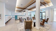 Located at 195 River Grove Way in West Palm Beach, Florida, this beautifully designed lobby serves as the welcoming heart of the community. The space showcases a refined coastal-modern aesthetic, defined by soaring ceilings accented with warm wood beam detailing, recessed lighting, and crisp white walls that create an open, airy atmosphere throughout.

The main lounge area is generously proportioned, furnished with a curated mix of upholstered wingback chairs, slatted wood accent chairs, and sleek dark side tables, all arranged atop layered vintage-style area rugs over large-format tile flooring. Bold, colorful artwork is displayed within custom wood-framed niches along one wall, adding a vibrant focal point to the seating area.

A dedicated mail center runs prominently through the space, featuring rows of white individual mailboxes trimmed in a signature teal accent — a detail that carries throughout the lobby's color palette. Lush potted plants, including fiddle-leaf figs, introduce a touch of greenery and warmth.

Large glass entry doors and floor-to-ceiling windows flood the space with natural light, offering views of swaying palm trees outside. Additional lounge seating near a bar or café area provides further gathering space, making this lobby a functional and welcoming environment for residents.