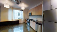 This contemporary single-floor space showcases a thoughtfully designed open-concept layout that blends functionality with modern urban aesthetics. The kitchen is a standout feature, outfitted with a full suite of stainless steel appliances including a gas range with oven, built-in microwave, refrigerator, and dishwasher. Rich dark granite countertops provide a striking contrast against the warm-toned flat-panel cabinetry that extends to the ceiling, offering generous storage throughout. A subtle tile backsplash adds texture and detail to the kitchen area, complementing the overall design palette. The polished concrete floors run throughout the space, lending an industrial character while remaining easy to maintain and highly durable. Large windows allow abundant natural light to pour in, creating a bright and airy atmosphere with views of the surrounding urban environment. A pendant light fixture anchors the open living area, while an industrial-style ceiling fan adds both function and a distinctive design element. The cohesive blend of raw materials, modern finishes, and efficient use of space gives this residence a sleek, loft-inspired character well-suited to contemporary urban living.