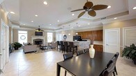 This single-floor space showcases an open-concept layout that effortlessly connects the kitchen, dining, and living areas into one cohesive and functional environment. The kitchen is anchored by rich, warm-toned cabinetry and a striking stone-accented island with bar seating, providing both a practical workspace and a casual gathering spot. Black appliances and pendant lighting add a refined contrast to the neutral palette. The dining area features a substantial dark-finished table and chairs, positioned to take full advantage of the open sightlines across the space. Flowing into the living area, the room is anchored by a classic fireplace with a white mantle surround, a wall-mounted television, and comfortable seating arranged atop a patterned area rug. Large ceiling fans with bronze finishes are installed throughout, contributing to both the aesthetic and the airflow of the space. Recessed lighting is thoughtfully distributed across the tray ceilings, which add architectural depth and definition to each zone. Light large-format tile flooring runs continuously throughout, enhancing the sense of spaciousness. Crown molding and neutral wall tones complete the polished, inviting ambiance of this well-appointed single-level space.