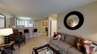 This single-floor residence features an open-concept layout that connects the living, dining, and kitchen areas into one cohesive space. The living area is anchored by a comfortable sofa accented with colorful throw pillows, a glass-topped coffee table, and a large round decorative mirror that adds a striking focal point to the neutral-toned walls. Plush wall-to-wall carpeting runs throughout, contributing to a warm, comfortable ambiance.

The kitchen is positioned along one side of the open floor plan and is delineated by a raised breakfast bar with seating for two, providing both a casual dining option and a natural visual boundary between cooking and living spaces. White cabinetry and appliances are visible from the main living area, keeping the palette light and cohesive.

Beyond the living space, a doorway leads to additional rooms, and a closet with a paneled white door is conveniently accessible. The textured ceiling and recessed lighting add subtle architectural detail throughout. The overall atmosphere is functional and welcoming, with a straightforward, practical design well-suited for everyday living.