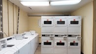 This shared laundry facility offers a practical and organized space designed to accommodate multiple users efficiently. The room is lined with a row of top-loading washing machines along one wall, each clearly accessible and ready for use. Opposite, a bank of stacked coin-operated dryers — numbered for easy identification — maximizes the available floor space while providing ample drying capacity. The units are neatly arranged and well-maintained, with red-numbered labels on each dryer for straightforward reference. Overhead fluorescent lighting illuminates the entire room evenly, ensuring a bright and functional environment. The walls feature a neutral, light-toned finish, and the flooring is a clean, low-maintenance hard surface. A visible ductwork run along the upper wall suggests dedicated ventilation to support proper airflow for the dryers. Informational signage is posted near the machines, offering guidance on proper machine usage. Overall, the laundry room is thoughtfully laid out to provide a convenient, no-frills space that prioritizes utility and ease of use for its occupants.